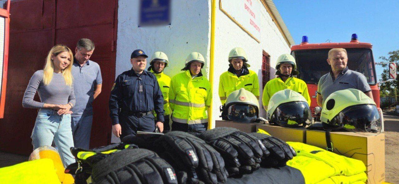 Група із восьми осіб, включно з жінкою у світлій смугастій кофті та джинсах (ліворуч), двох чоловіків у світлих сорочках, чоловіком у пожежній формі та чотирма пожежниками у жовтому захисному одязі та шоломах, які стоять на вулиці біля будівлі. На передньому плані на столі розкладено нове пожежне екіпірування: захисні шоломи (каски), чорні рукавички та яскраво-жовті куртки.