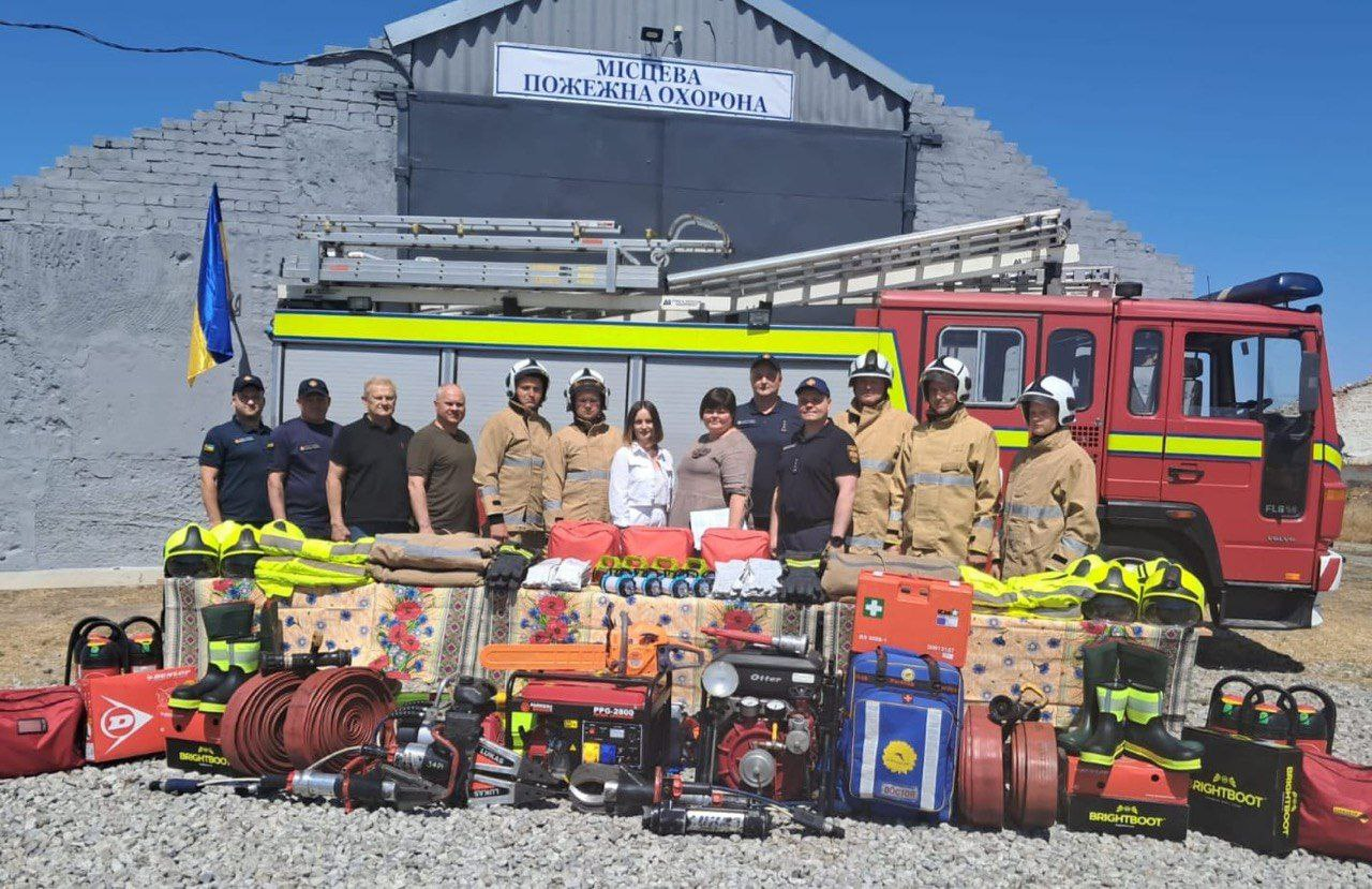 Група людей, більшість з яких – чоловіки в пожежній формі та шоломах, а також дві жінки в цивільному одязі, що стоять перед червоною пожежною машиною. Вони стоять на тлі сірої будівлі, на фасаді якої видно напис: МІСЦЕВА ПОЖЕЖНА ОХОРОНА. Перед групою та машиною на землі, вкритій ковдрами, розміщене велике різноманіття нового пожежного та рятувального обладнання, включаючи насоси, бензопили, вогнегасники, рукави, чоботи (RIGHTBOOT та BRIGHTBOOT), аптечки та інші інструменти. Зліва також видно прапор України.