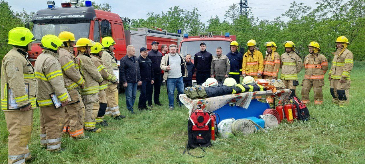 Група чоловіків, частина з яких одягнена у пожежну форму (жовто-зелені захисні костюми та шоломи), стоять на трав'янистій галявині біля червоної пожежної машини. Інші чоловіки, одягнені у цивільний та військовий одяг, стоять посередині. У центрі композиції на землі розкладене пожежне обладнання, включаючи вогнегасники, пожежні рукави та манекен, одягнений у захисну екіпіровку і лежачий на ношах. Сцена виглядає як інструктаж або навчання рятувальників.