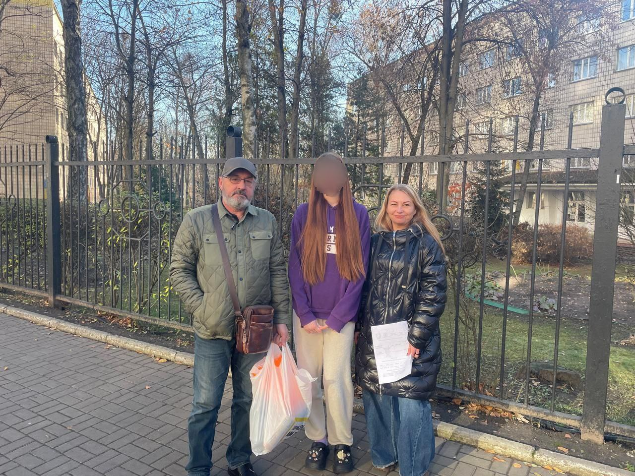 Чоловік у зеленій куртці, дівчина у фіолетовому світшоті та жінка у чорній куртці стоять на вулиці перед металевим парканом і деревами. Чоловік тримає білий пакет, жінка — документи. Обличчя дівчини приховане мозаїкою для збереження приватності.