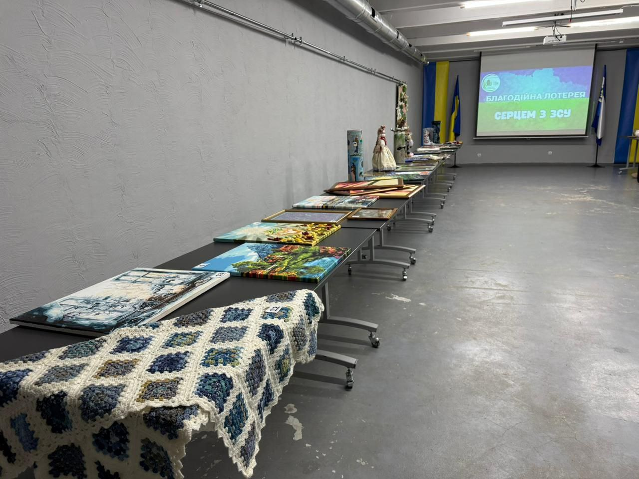 Довгий ряд столів з картинами та виробами на виставці, включно з в'язаним покривалом. На задньому плані видно екран із написом: БЛАГОДІЙНА ЛОТЕРЕЯ СЕРЦЕМ З ЗСУ та українські прапори.