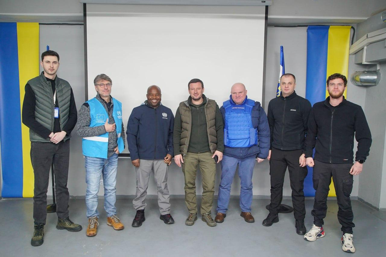 Група з семи чоловіків стоїть у ряд, позуючи для фотографії. Двоє з них одягнені у жилети гуманітарних організацій (зокрема ООН/OCHA). На задньому плані видно екран, а також вертикально розташовані українські прапори.
