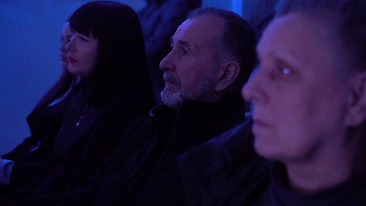 Група людей, включаючи чоловіка з бородою в центрі, сидять в темному приміщенні, освітленому синім світлом, дивлячись уперед. Жінка з темним волоссям і яскраво-червоною помадою сидить ліворуч від чоловіка.