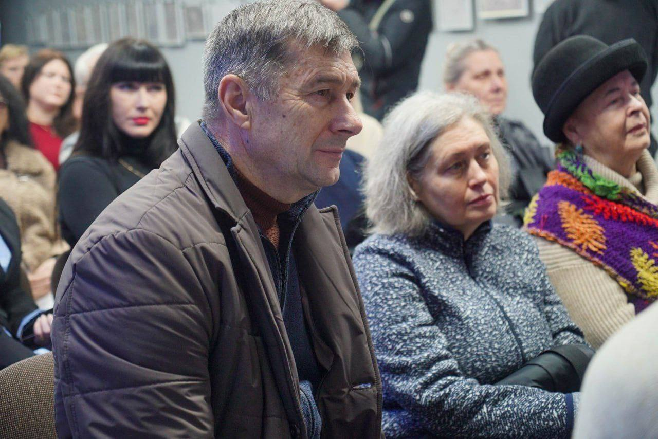 Чоловік у коричневій стьобаній куртці сидить на передньому плані, поруч із ним жінка зі світлим кучерявим волоссям у синій стьобаній куртці, вони дивляться вбік. На задньому плані видно інших людей, які слухають.