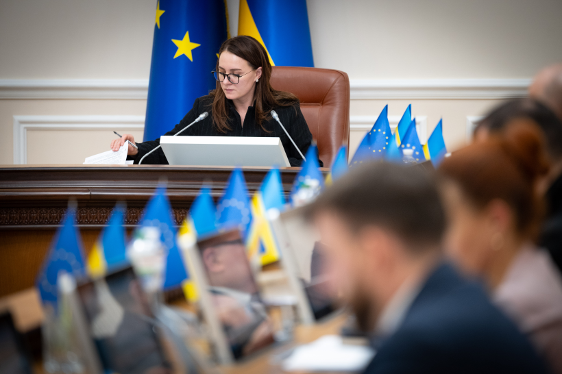 Жінка в окулярах переглядає документи за столом президії, оточеним маленькими прапорцями України та ЄС під час наради.