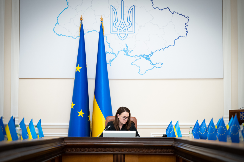 Жінка за великим столом для нарад на фоні прапорів України, ЄС та карти України з гербом.