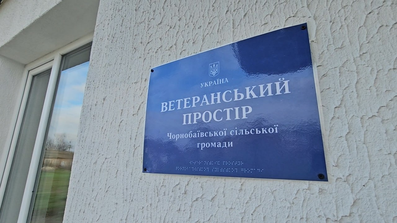 Синя табличка на світлій стіні з написом: Ветеранський простір Чорнобаївської сільської громади та шрифтом Брайля.
