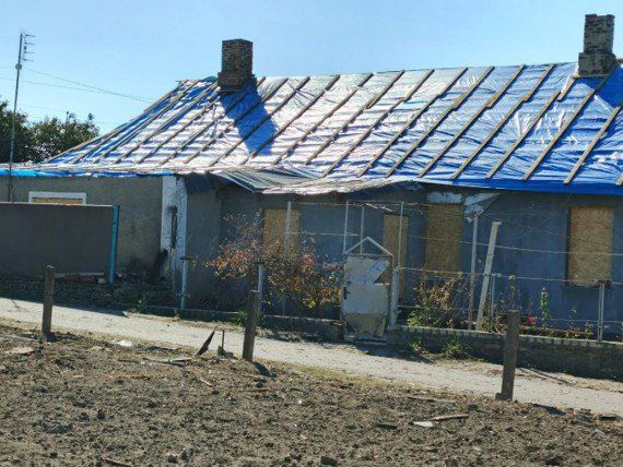 Одноповерховий приватний будинок, пошкоджений внаслідок бойових дій. Дах будівлі повністю накритий захисною блакитною плівкою, закріпленою дерев'яними рейками, а вікна забиті листами ОСБ-плит.