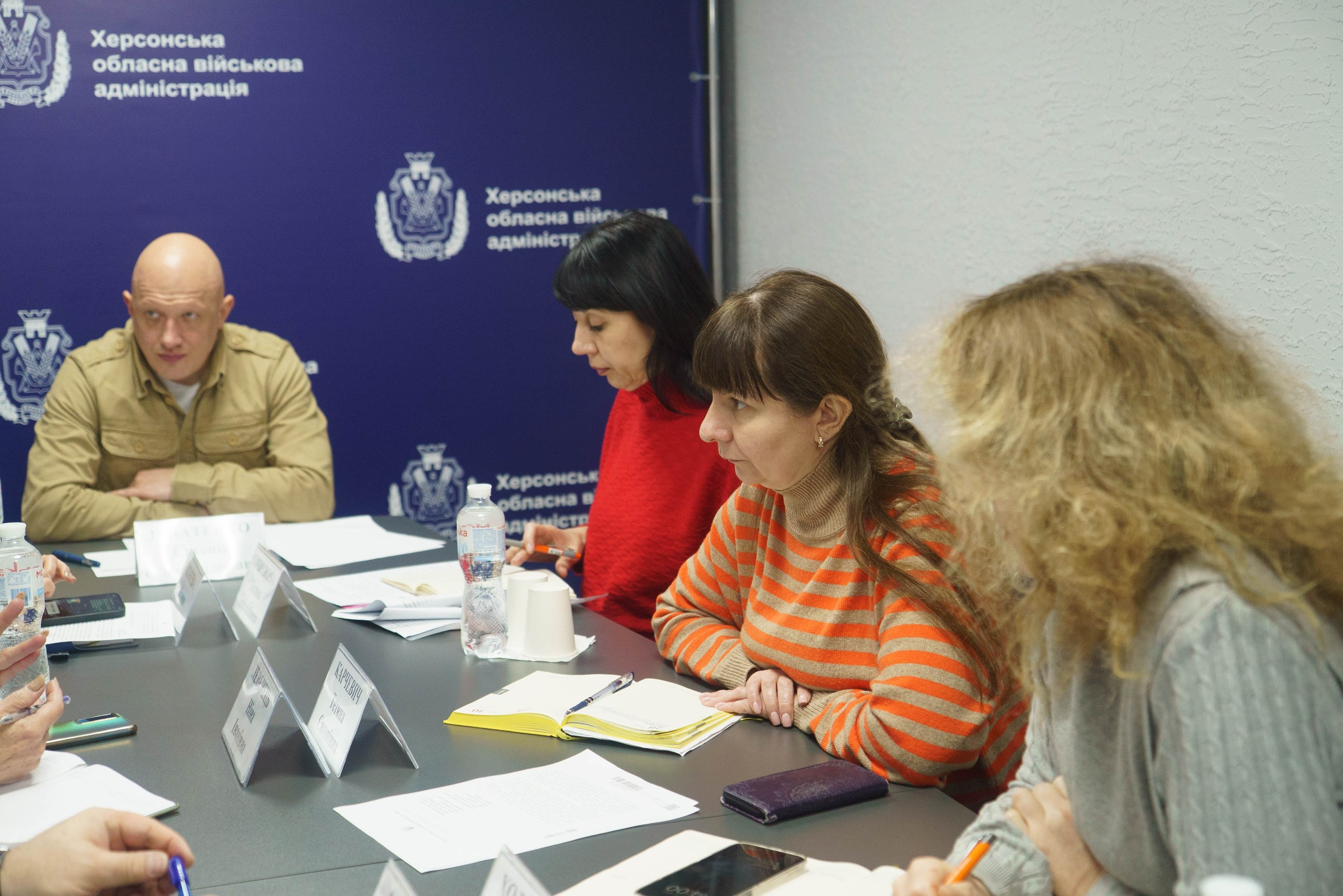 Учасники офіційної зустрічі сидять за робочим столом, занотовуючи інформацію під час обговорення. На фоні праворуч розміщено синій банер із символікою та написом: Херсонська обласна військова адміністрація.