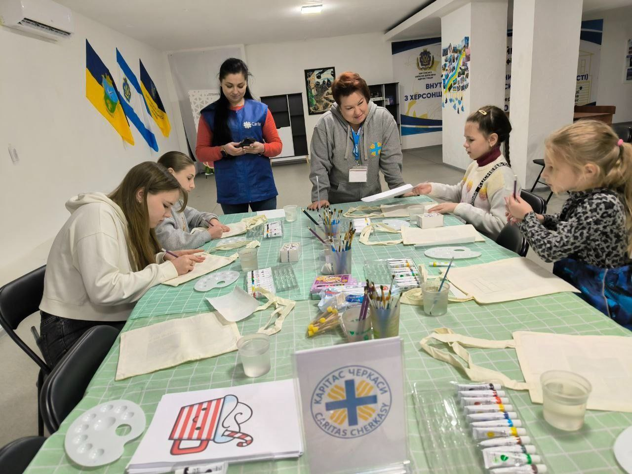 Творче заняття для дітей у світлому приміщенні. Група дівчат сидить за великим столом, застеленим зеленою скатертиною, і розмальовує екосумки. На столі розкладені фарби, пензлики, палітри та зразки малюнків. Поруч із дітьми стоять двоє дорослих жінок у жилетках із символікою благодійних організацій, які допомагають у процесі. На задньому плані видно прапори та інформаційні стенди.