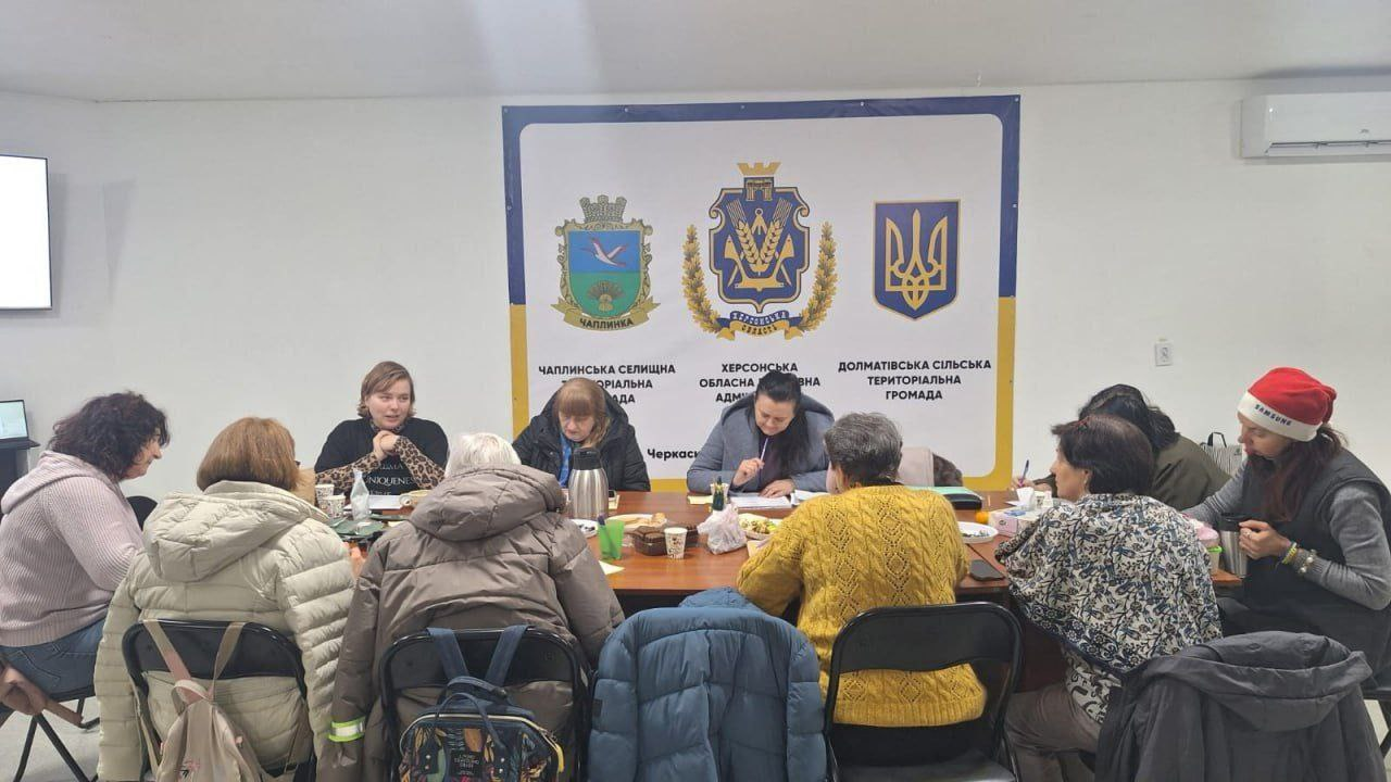 Робоча зустріч представників громад Херсонщини. Група жінок сидить за круглим столом у світлому приміщенні під час обговорення. На фоні розміщено банер із гербами Чаплинської селищної, Херсонської обласної та Долматівської сільської територіальних громад. Учасниці роблять нотатки, на столі стоять чашки та особисті речі.