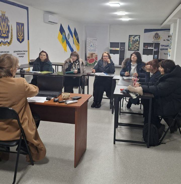 Група жінок бере участь у тренінгу, сидячи за столами в офісному приміщенні. На стінах видно державну символіку, прапори та інформаційні стенди. Учасниці зосереджено слухають та роблять записи.