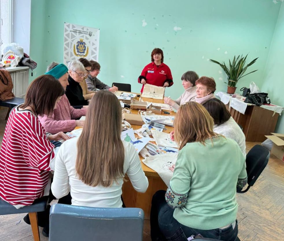 Група жінок сидить навколо великого столу у світлій залі та займається розписом екосумок. На фоні стоїть білий інформаційний банер Херсонської обласної державної адміністрації, а кімната прикрашена кімнатними рослинами.