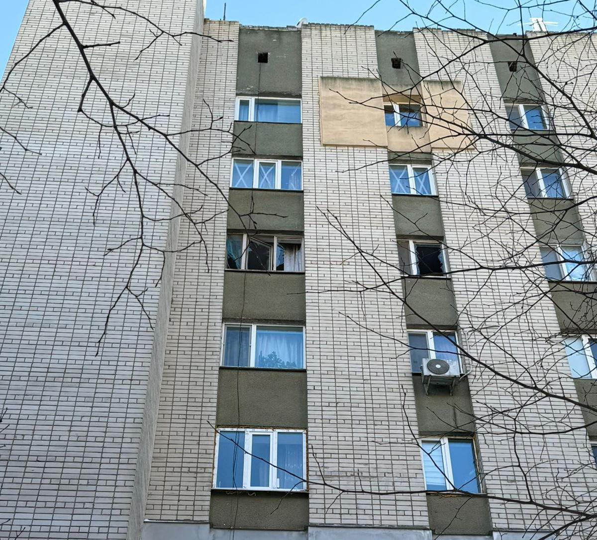 Фасад багатоповерхового цегляного будинку з пошкодженим склінням. У кількох вікнах на середніх поверхах вибите скло, видно темні порожнечі віконних рам. Перед будівлею — голе гілля дерев без листя на тлі ясного неба.