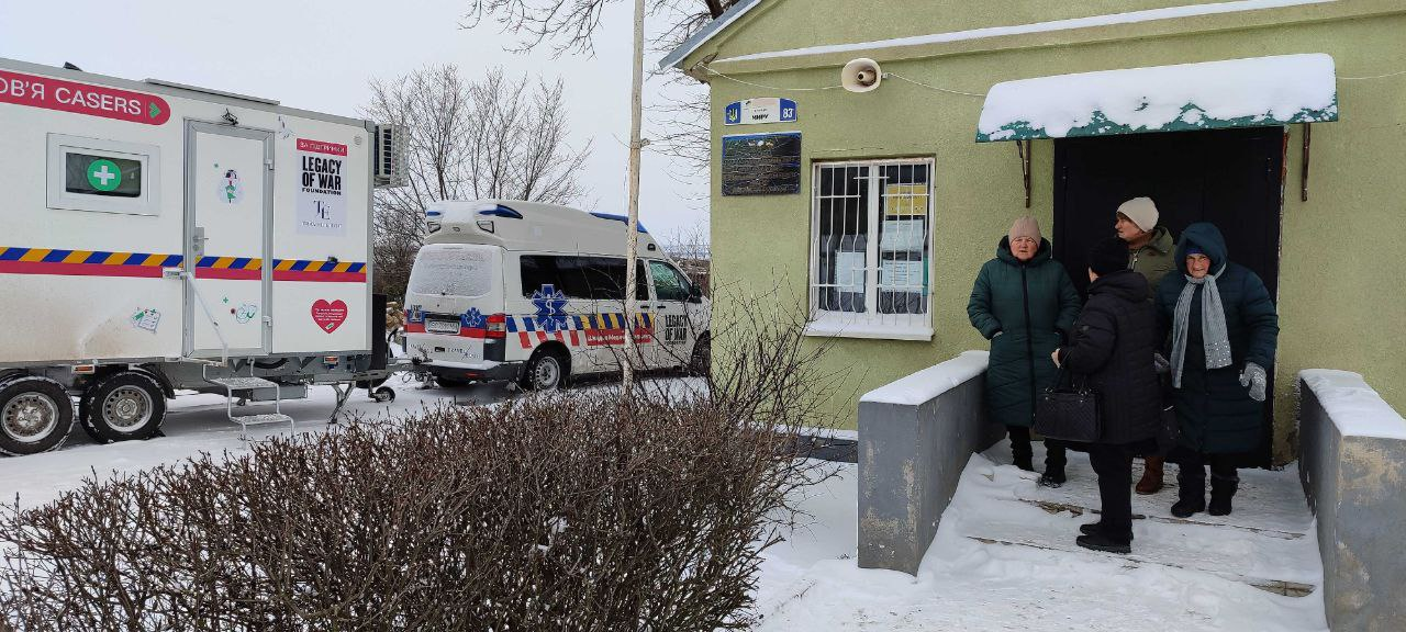 Світлина мобільного медичного пункту та реанімобіля організації «CASERS». На засніженому подвір'ї біля входу до будівлі стоїть група людей літнього віку, очікуючи на прийом або допомогу.