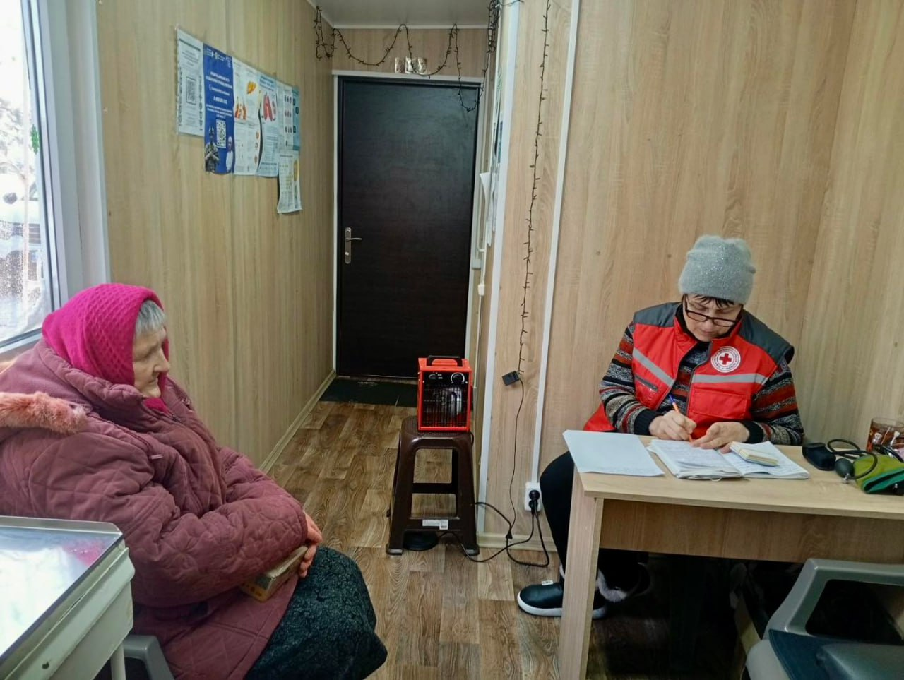 Працівниця гуманітарної організації у фірмовому жилеті заповнює документи за столом під час прийому літньої жінки в облаштованому приміщенні. На стіні розміщені інформаційні плакати, а поруч стоїть портативний обігрівач.