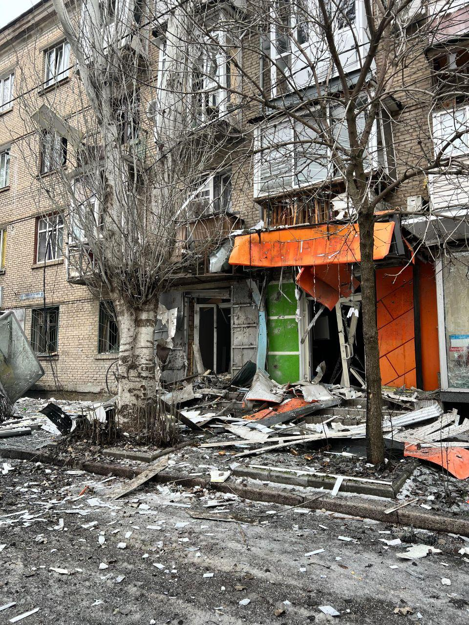 Наслідки обстрілу житлового будинку: зруйнований фасад першого поверху з понівеченими вхідними групами магазинів, розбитим склом та купами будівельного сміття на тротуарі перед цегляною багатоповерхівкою.