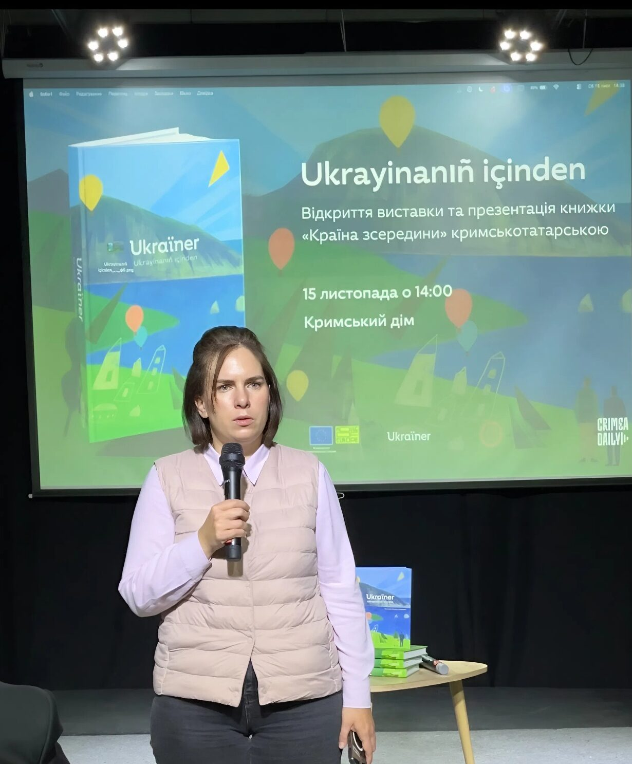 Жінка виступає з мікрофоном на фоні великого екрана з анонсом презентації книги «Ukrayinanıñ içinden» («Країна зсередини») кримськотатарською мовою. На передньому плані видно стіл із примірниками видання.