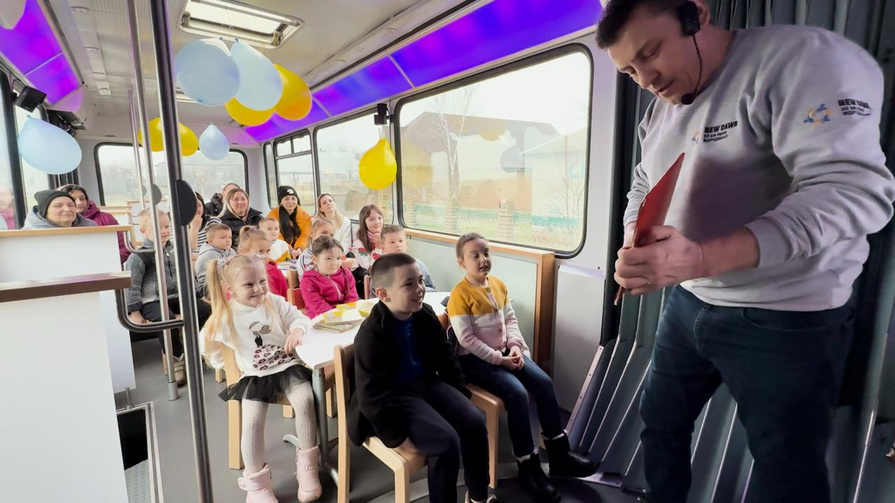 Аніматор у світшоті з гарнітурою проводить розважально-освітнє заняття для групи дітей та дорослих у салоні спеціально облаштованого автобуса. Інтер’єр прикрашений повітряними кульками, а діти зацікавлено спостерігають за ведучим.