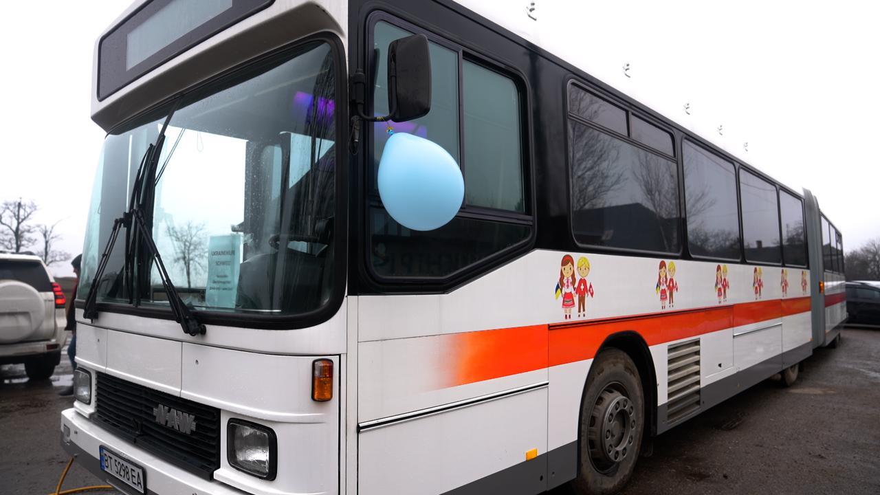 Білий пасажирський автобус із помаранчевою смугою вздовж борту та малюнками дітей у національному вбранні. До дзеркала прикріплена блакитна повітряна кулька.