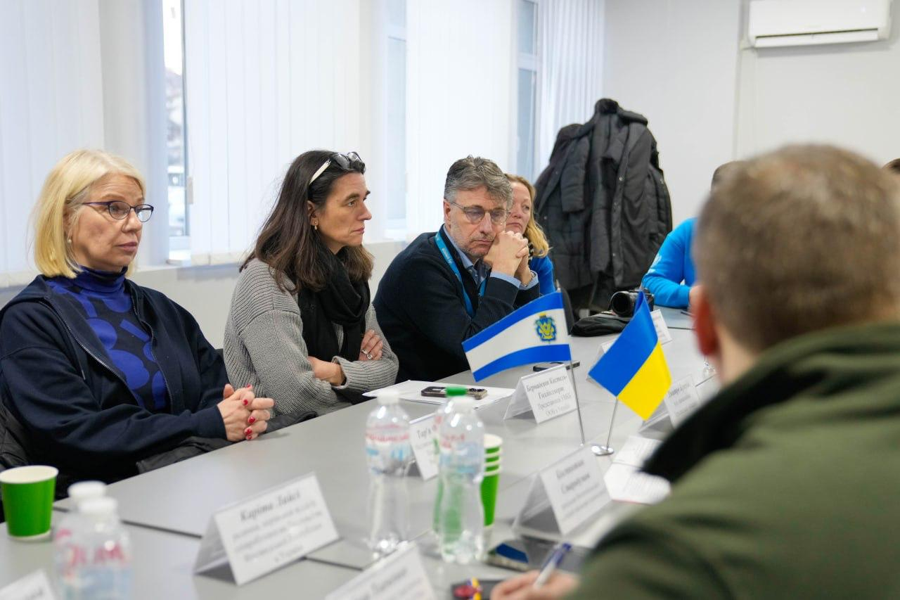 Дві жінки та чоловік сидять за столом під час ділової зустрічі. Вони одягнені в повсякденний офісний одяг і уважно слухають доповідача, чию потилицю видно на передньому плані. На столі стоять прапорці, вода в пластикових пляшках та паперові стаканчики.