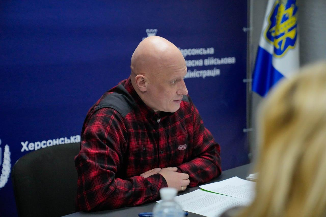 Портрет чоловіка у червоній клітчастій сорочці, який сидить за столом під час наради. Він уважно дивиться у бік співрозмовників. На фоні видно синій банер з написом «Херсонська обласна військова адміністрація» та прапор Херсонської області.