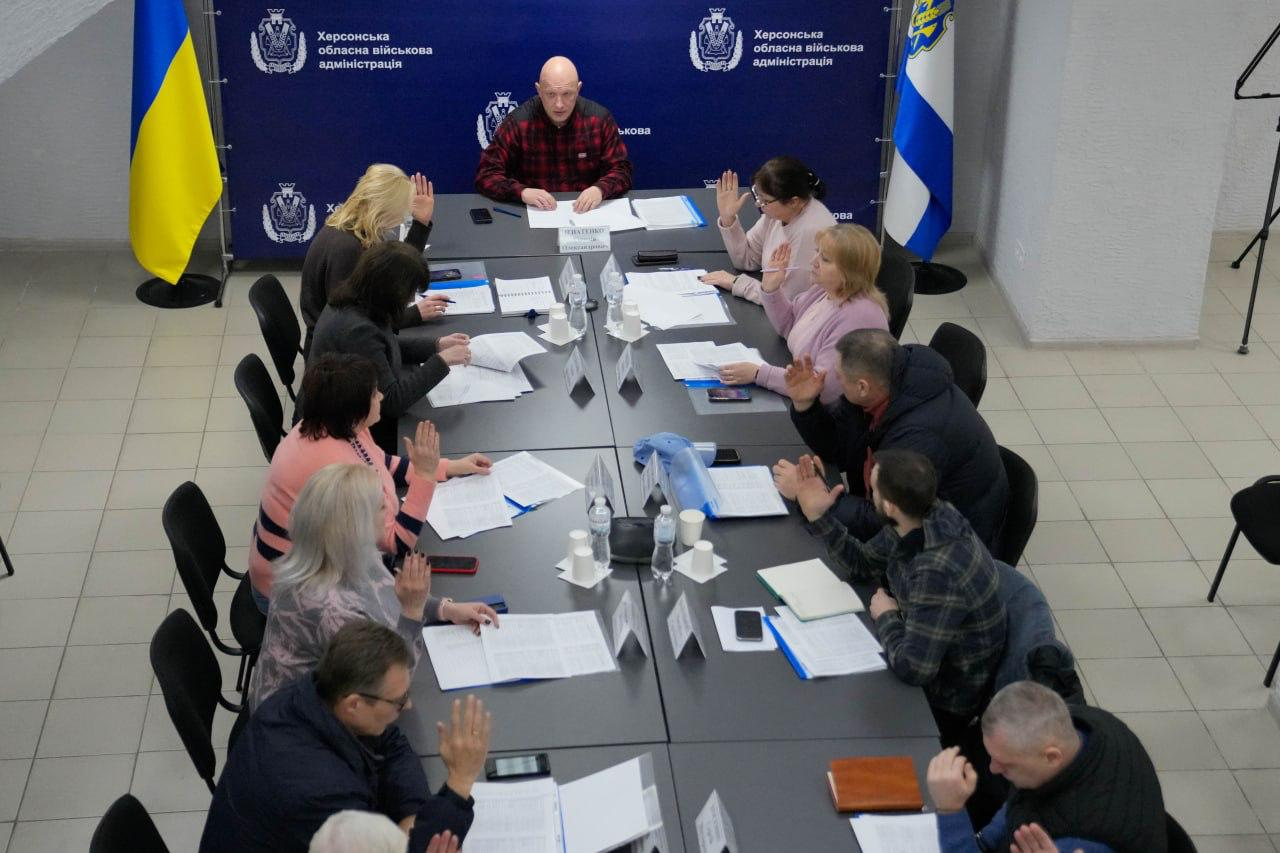Учасники офіційної зустрічі в Херсонській ОВА сидять за довгим столом і піднімають руки під час голосування. У президії засідає чоловік у червоній сорочці в клітинку. На фоні розташований синій банер з логотипами установи та державні прапори України й Херсонської області.