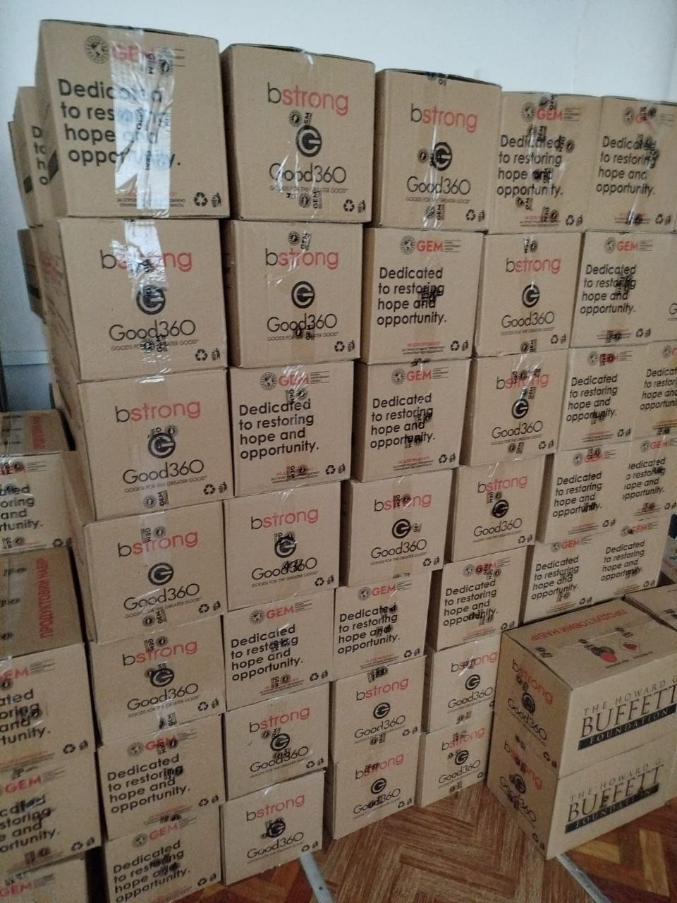 Стіна з численних картонних коробок гуманітарної допомоги, складених одна на одну. На пакуваннях надруковано логотипи організацій «bstrong», «Good360» та «GEM» із гаслом «Dedicated to restoring hope and opportunity». Праворуч внизу видно частину коробки з написом «The Howard G. Buffett Foundation».