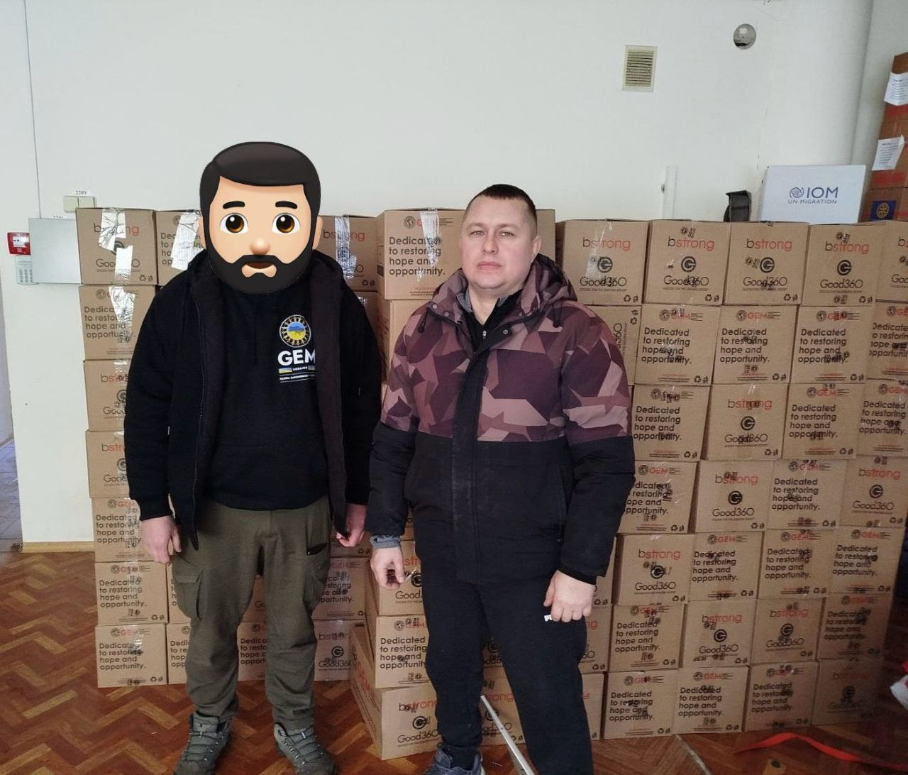 Двоє чоловіків стоять у приміщенні на фоні численних картонних коробок із гуманітарною допомогою. На коробках розміщені логотипи організацій «GEM», «bstrong» та «Good360». Один із чоловіків, обличчя якого приховане, одягнений у чорну худі з емблемою «GEM», інший — у камуфляжну куртку. На задньому плані також видно пакування з логотипом «IOM UN Migration».