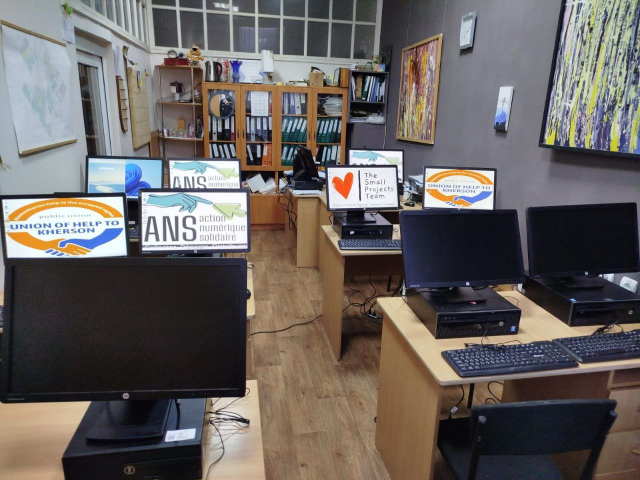 Робочий кабінет з декількома комп'ютерними станціями на дерев'яних столах. На моніторах відображені логотипи благодійних організацій: «The Small Projects Team», «Union of Help to Kherson» та «ANS». У фоні видно шафу з папками для документів та картини на стінах.