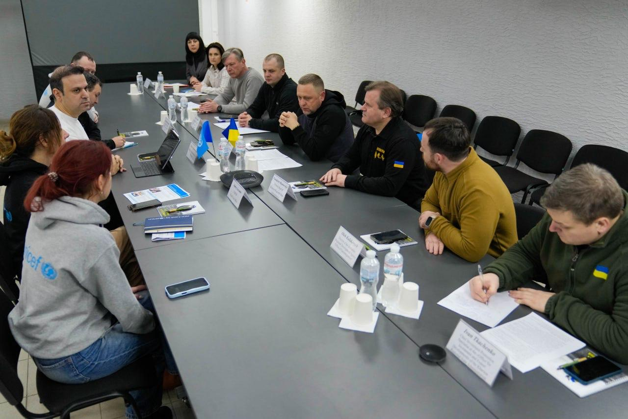 Ответ Gemini Ось короткий альтернативний текст для третього фото: Група людей сидить за довгим столом під час офіційної зустрічі. Учасники обговорюють робочі питання, перед ними лежать документи, блокноти та стоять пляшки з водою. На столі встановлено маленькі прапори України та ООН, а також таблички з іменами. Деякі присутні одягнені в худі з логотипом ЮНІСЕФ.