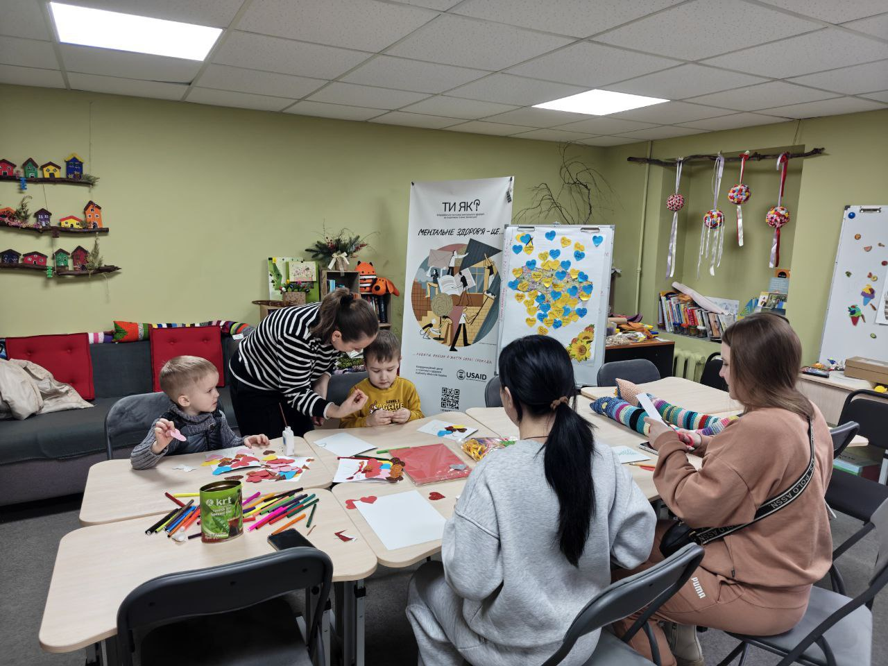 Дорослі та діти займаються творчістю за великим столом у світлій кімнаті. Вони вирізають аплікації та малюють. На фоні видно інформаційний стенд із мапою України, прикрашеною паперовими серцями, та полиці з яскравим декором.