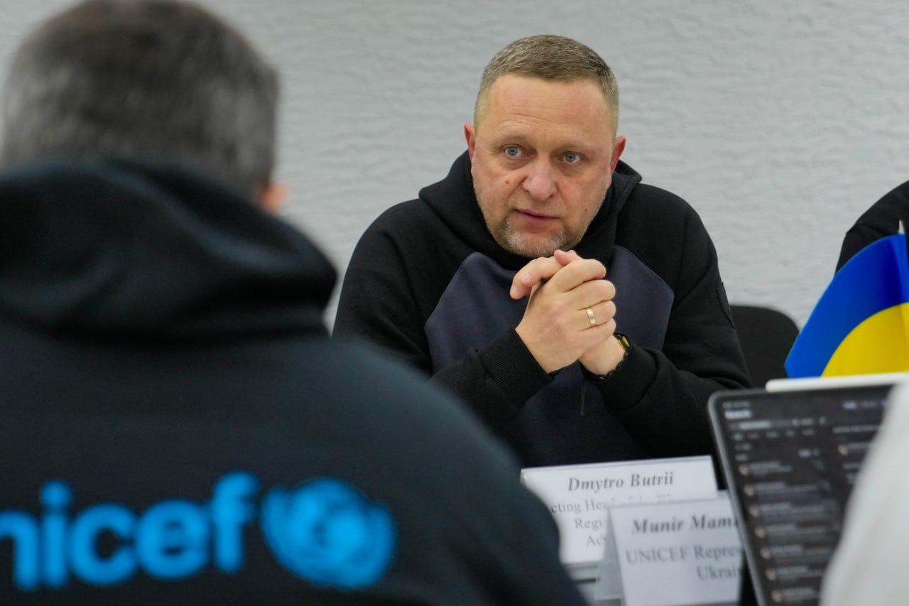 Чоловік у темному одязі сидить за робочим столом, склавши руки перед собою, та уважно слухає співрозмовника. Перед ним стоїть табличка з ім'ям, а на передньому плані видно спину іншого учасника зустрічі в худі з офіційним логотипом міжнародної організації.