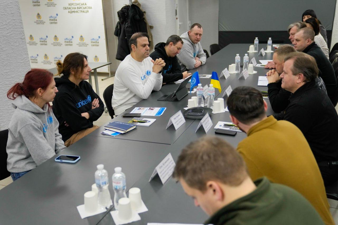 Група людей під час офіційної зустрічі за довгим столом у конференц-залі. Учасники з одного боку столу одягнені в білі та сірі худі з логотипом міжнародної організації, а присутні навпроти — у темний повсякденний одяг. На столі розміщені таблички з іменами, ноутбуки, документи та маленькі прапорці України та ООН.