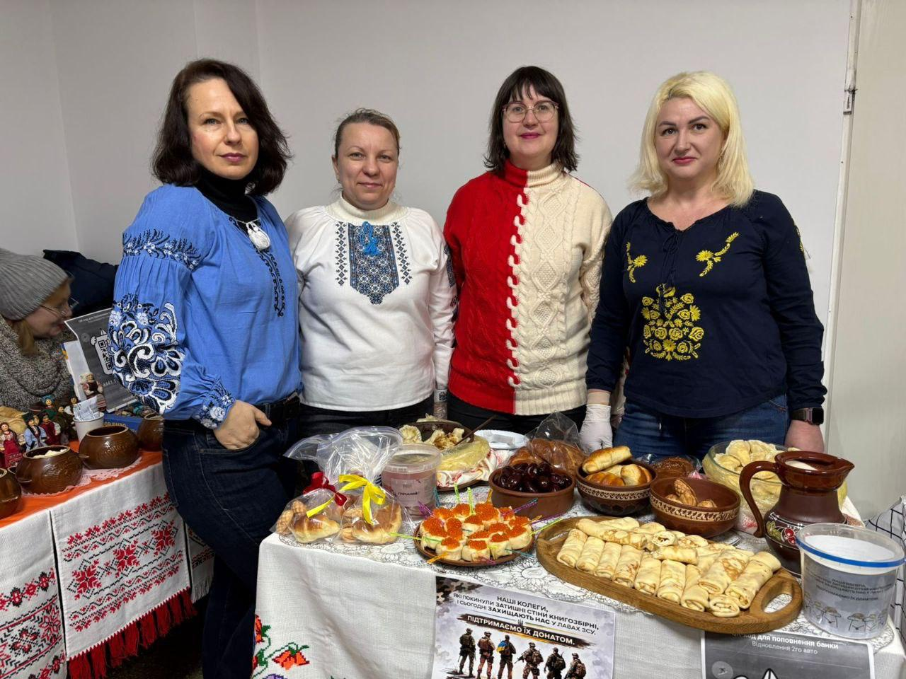 Чотири жінки у традиційному українському вбранні та вишиванках стоять за столом, накритим для благодійного ярмарку. На столі представлені різноманітні домашні страви: млинці, випічка, вареники та закуски. Попереду розміщено плакат із закликом підтримати колег, які захищають Україну в лавах ЗСУ.