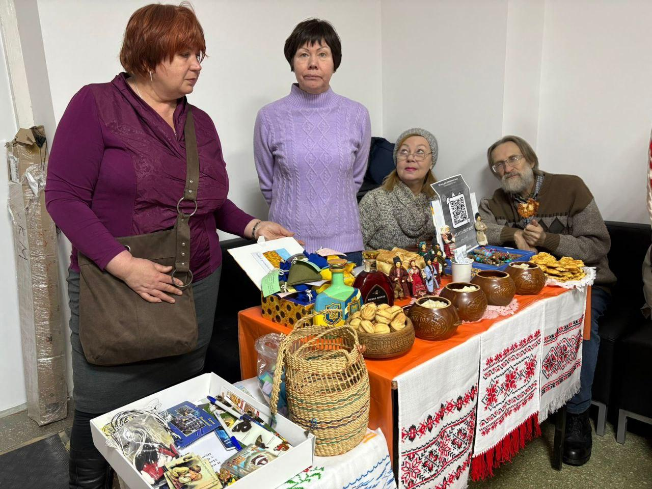 Дві жінки стоять біля столу з благодійними лотами та частуваннями. На столі, вкритому вишитим рушником, представлені сувеніри, патріотична атрибутика, випічка та глиняний посуд. Позаду сидять ще двоє учасників заходу, один із яких тримає частування, а поруч розміщено QR-код для збору коштів.