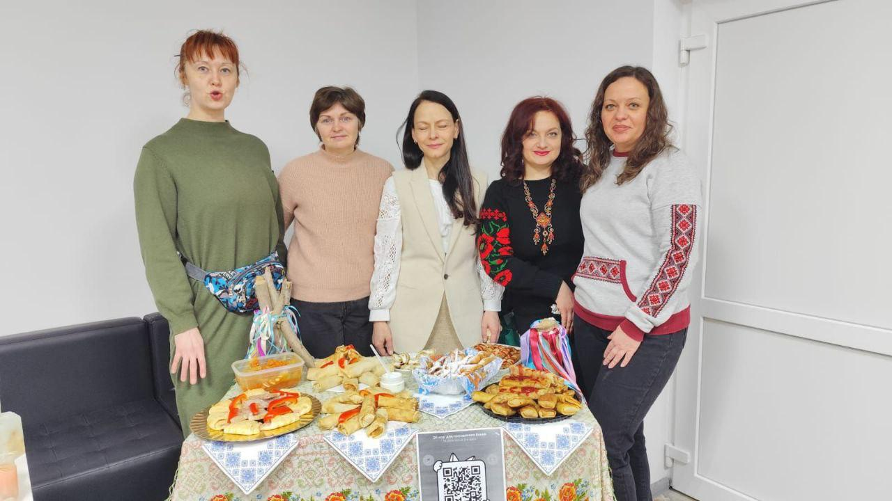 П'ять жінок стоять за святковим столом під час благодійного заходу. Деякі з них одягнені у вишиванки або одяг з етнічними мотивами. На столі, застеленому скатертиною з традиційним орнаментом, представлено різноманітну випічку та страви. Попереду розміщено оголошення з QR-кодом для збору коштів.