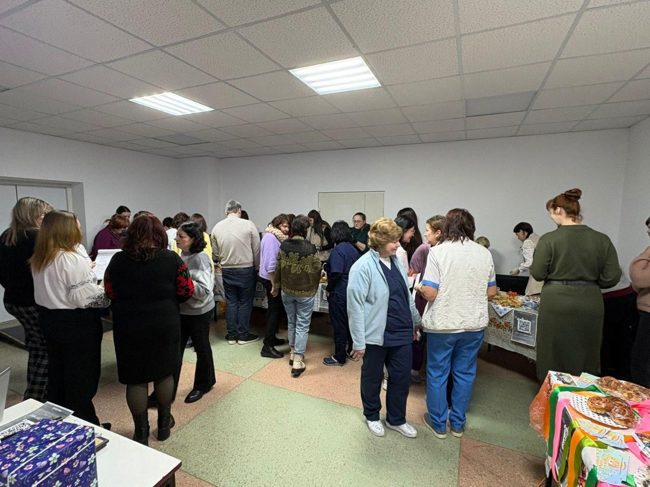 Велике скупчення людей у просторому світлому приміщенні під час проведення заходу. Учасники спілкуються між собою, стоять біля столів із частуваннями та патріотичною атрибутикою. Атмосфера події виглядає жвавою та робочою.