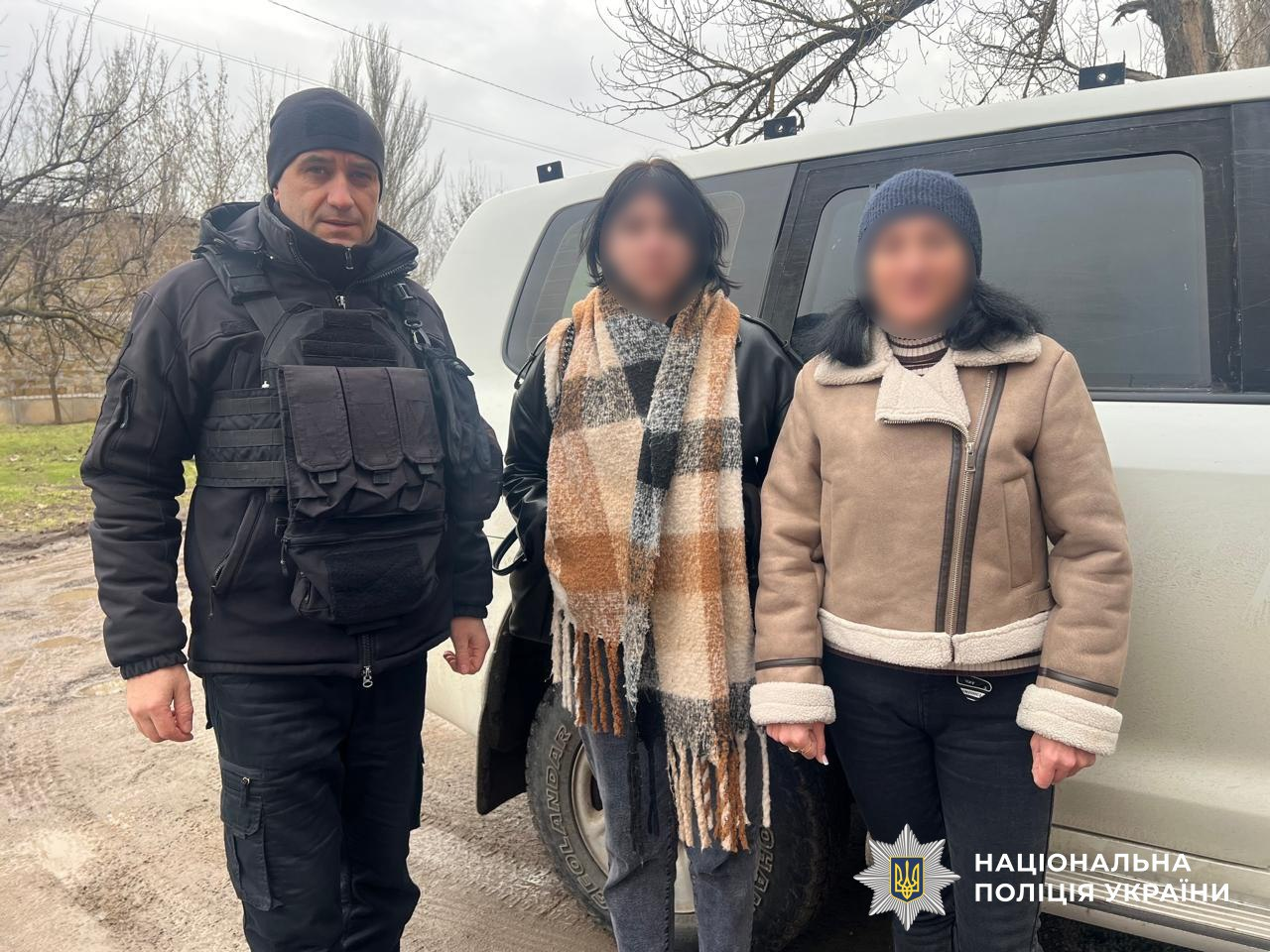 На фото зображено правоохоронця у повному захисному спорядженні та чорній формі, який стоїть поруч із двома жінками біля білого автомобіля. Обличчя цивільних осіб розмиті з міркувань приватності. У нижньому правому куті розміщено логотип та напис «Національна поліція України».