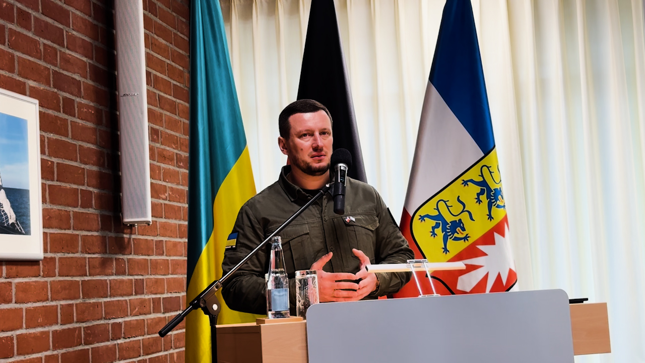 Чоловік у тактичному одязі виступає за трибуною з мікрофоном на фоні прапорів України та іноземного регіону. Він стоїть у приміщенні з цегляною стіною під час офіційного заходу.