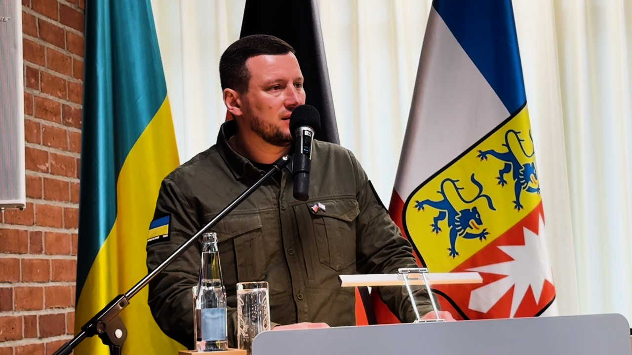 Чоловік у тактичній сорочці з мікрофоном виступає за трибуною. Він стоїть на фоні цегляної стіни та двох прапорів — України та землі Шлезвіг-Гольштейн (Німеччина).