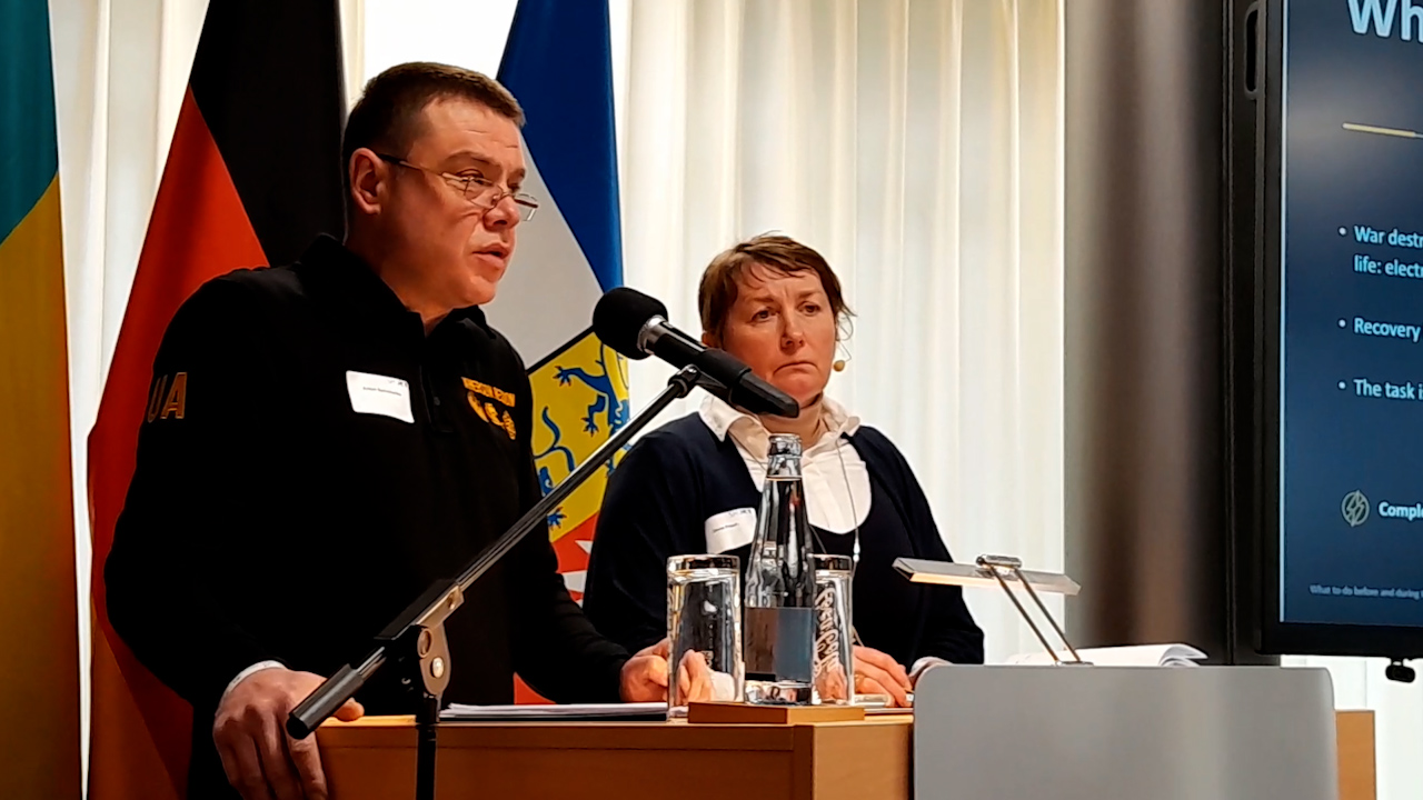 Чоловік в окулярах та чорній кофті з написом «UA» виступає з промовою за трибуною. Поруч із ним сидить жінка, яка уважно слухає. На фоні видно прапори та край екрана з презентацією.