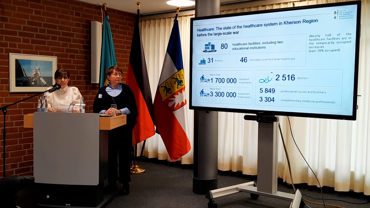 Жінка у білій блузі виступає за трибуною, поруч із нею стоїть інша жінка у темній кофті та синій сорочці. На великому екрані праворуч відображена презентація про стан медичної системи Херсонщини. У залі присутні державні прапори та цегляна стіна з картиною.