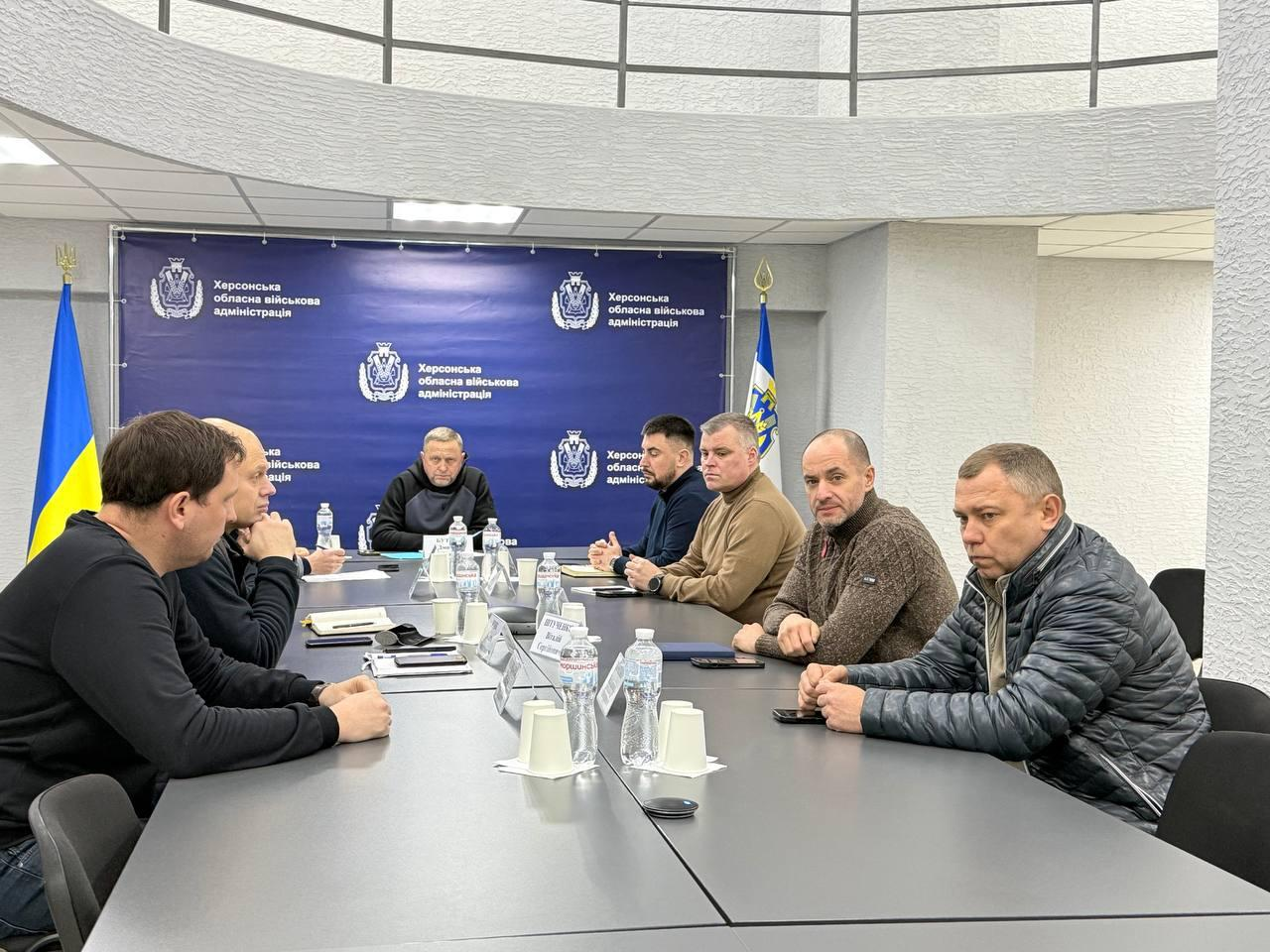 Група чоловіків у цивільному та мілітарі-одязі проводить робочу зустріч за великим столом у світлому кабінеті. На стіні за ними — синій банер із гербом та написом «Херсонська обласна військова адміністрація». По обидва боки від банера стоять державні прапори.