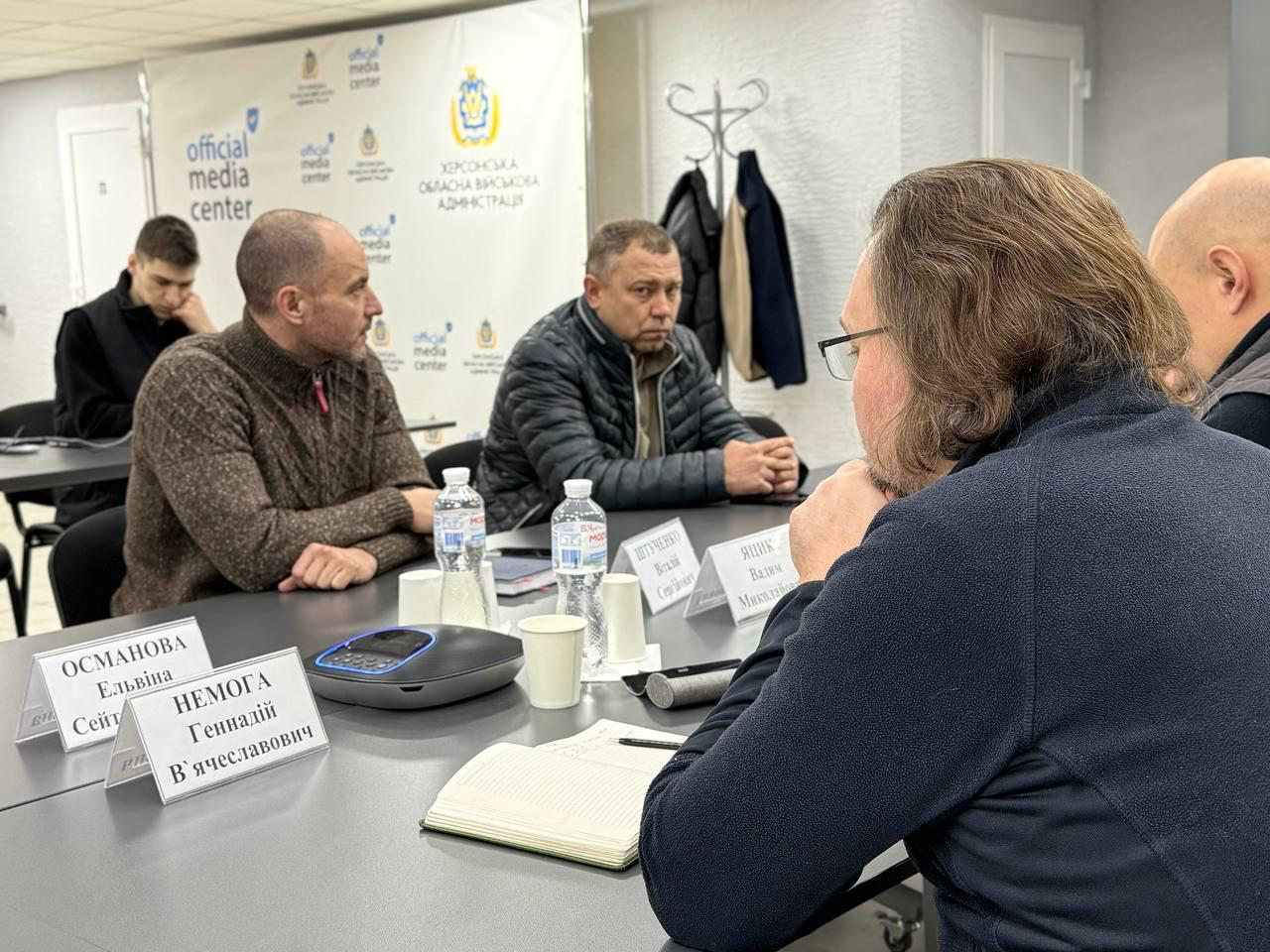 Група чоловіків сидить за робочим столом під час наради. На фоні видно світлий банер із логотипом «official media center» та гербом Херсонської обласної військової адміністрації.