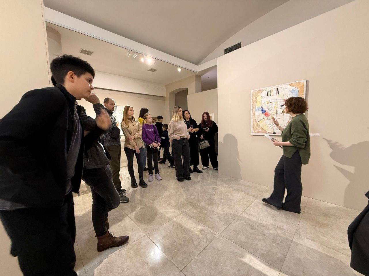 Група людей у залі художнього музею слухає екскурсовода, яка розповідає про картину, закріплену на стіні. На абстрактному полотні зображено стилізоване обличчя. Учасники заходу — дорослі та діти в повсякденному одязі — уважно спостерігають за презентацією.