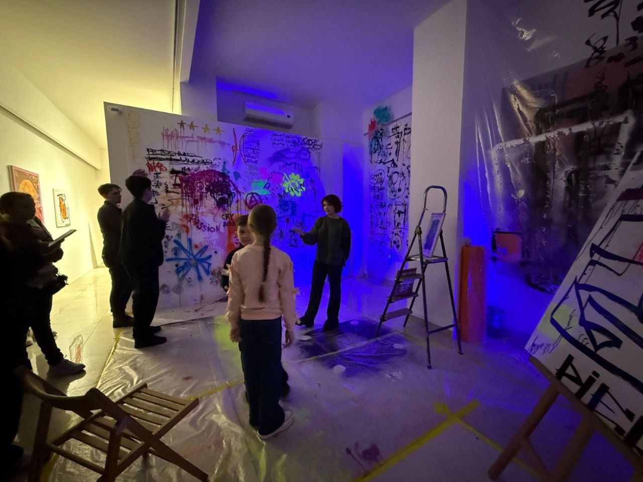 Екскурсовод та група відвідувачів у залі музею перед великою стіною, що розписана різнокольоровими графіті та написами. Приміщення підсвічене синім неоновим світлом, на підлозі розстелена захисна плівка, а поруч стоїть драбина та мольберт із картиною.
