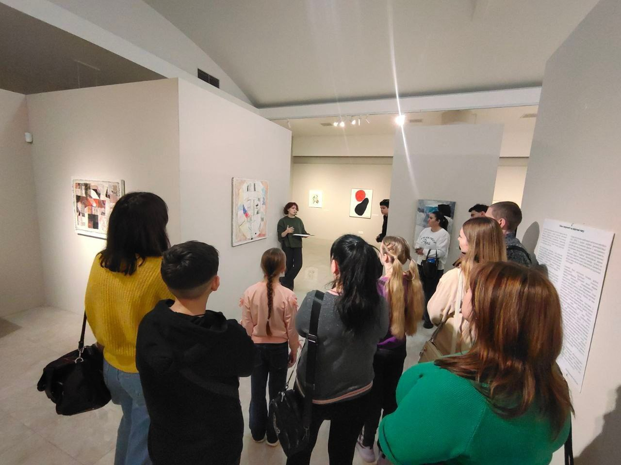 Група відвідувачів, серед яких дорослі та діти, стоять у музейній залі та слухають екскурсовода, яка проводить презентацію біля картини. На стінах розвішані полотна в стилі сучасного мистецтва, а праворуч розміщений інформаційний стенд із текстом.