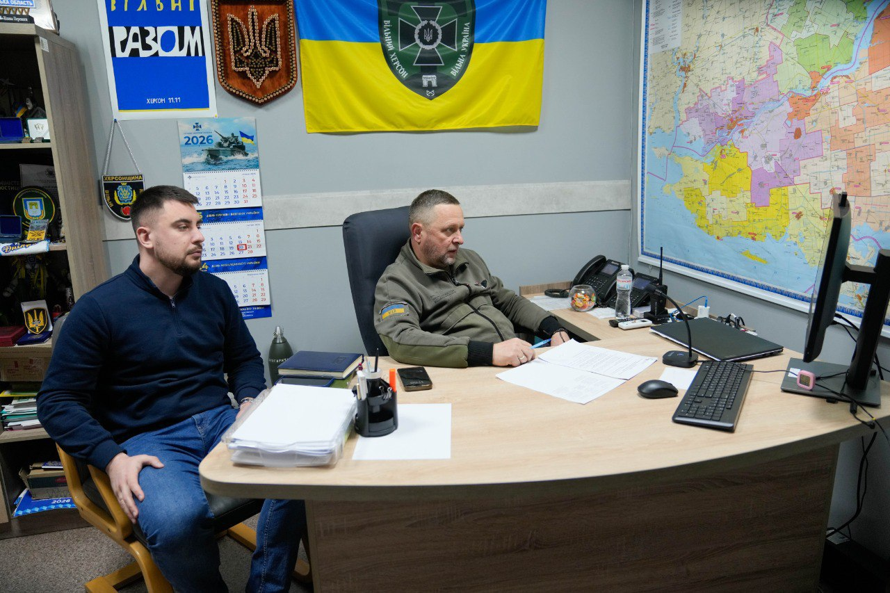 Двоє чоловіків сидять за робочим столом в офісному приміщенні. На стіні за ними розміщено Державний Прапор України з емблемою, дерев’яний герб та карту Херсонської області. На столі знаходяться документи, комп’ютерна техніка та засоби зв’язку.