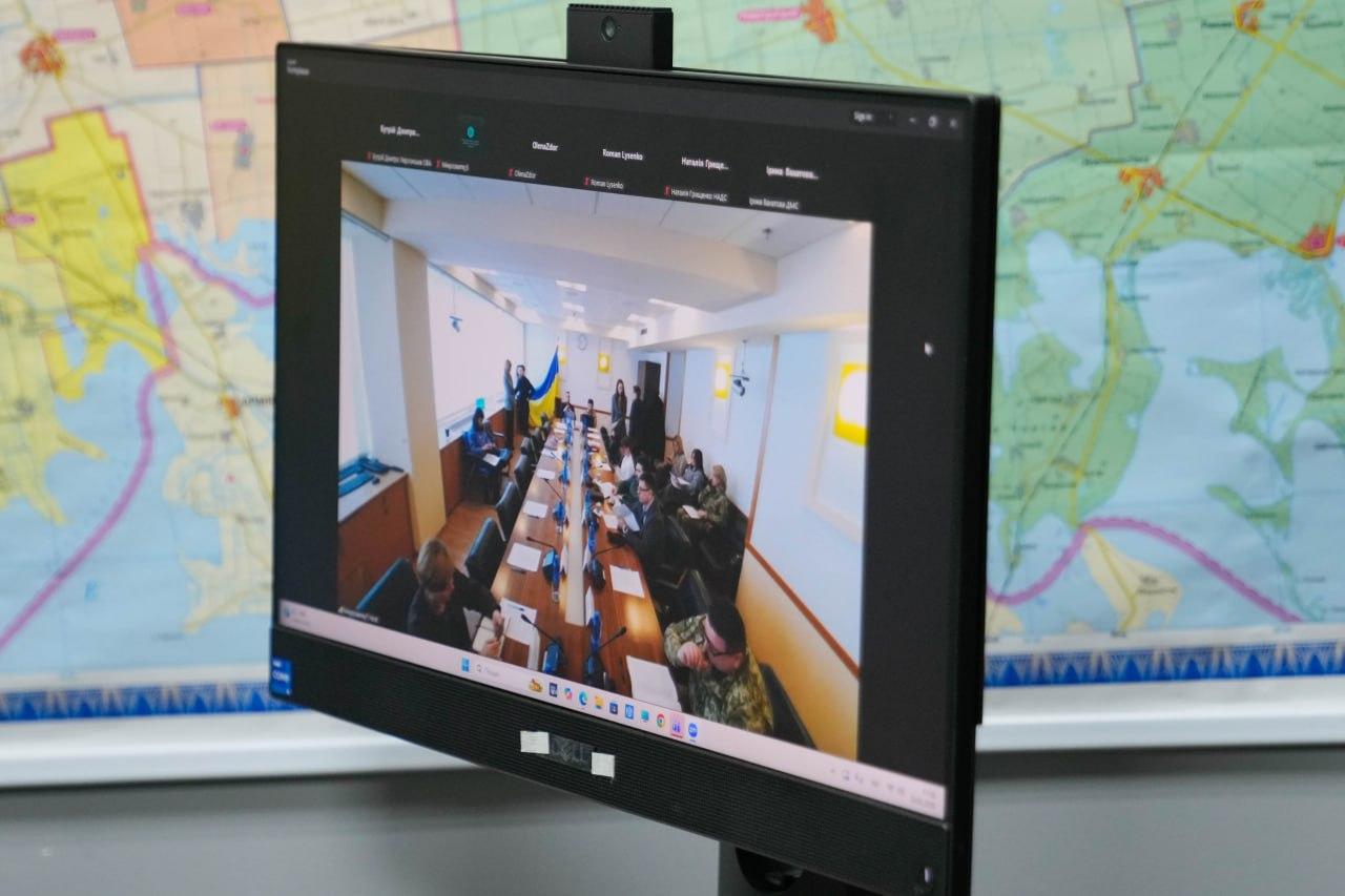 Монітор комп'ютера, на якому відображено вікно онлайн-конференції із прямою трансляцією із засідання у великій залі. На задньому плані за монітором видно частину настінної карти Херсонської області.